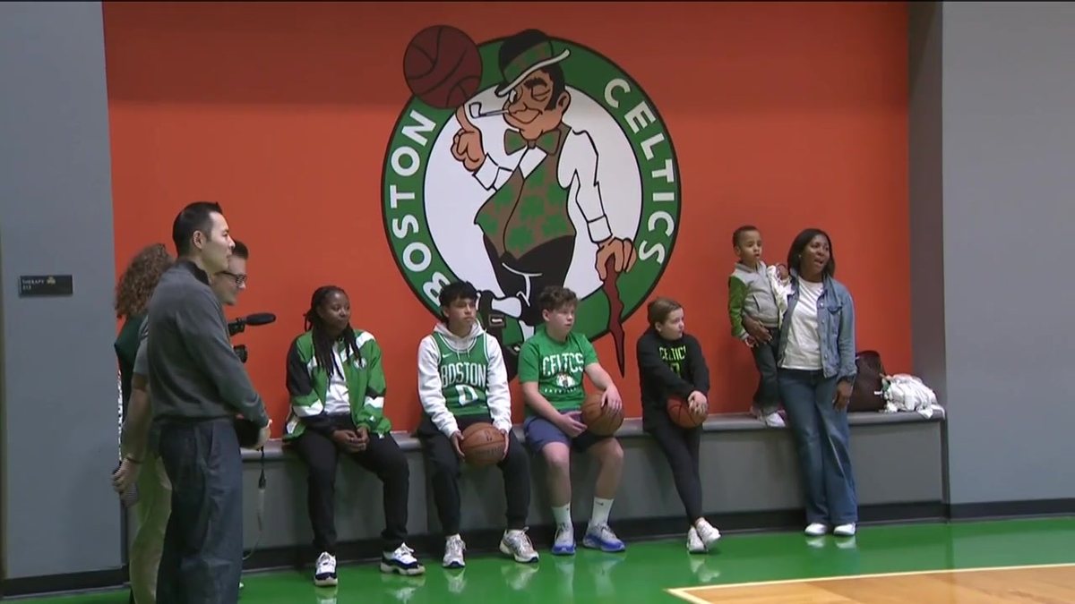 Kids in Celtics jerseys and jackets sit on a bench holding basketballs under a large Celtics logo at the team's practice court.