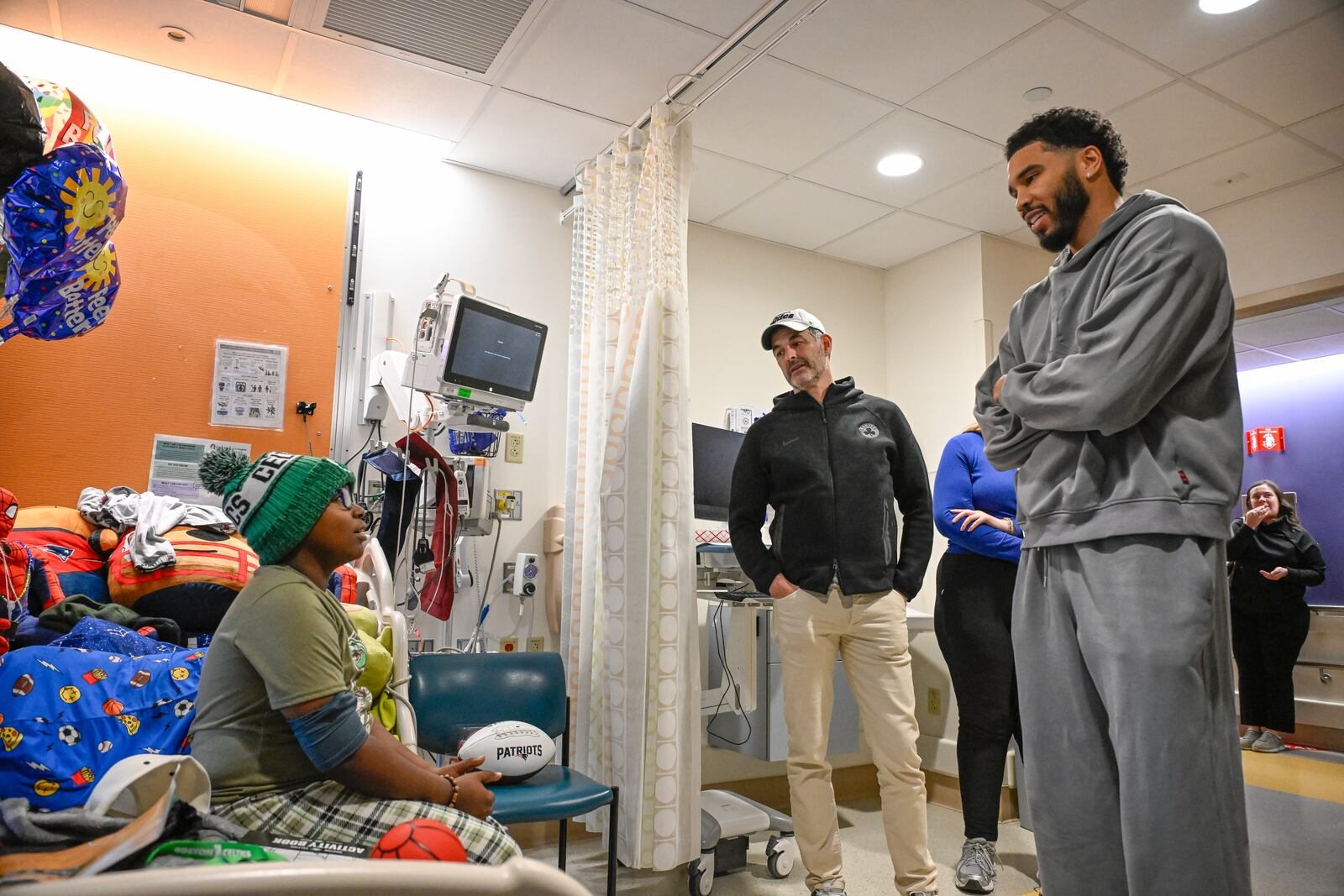 Jayson Tatum, in a gray sweatsuit, and Celtics owner Bill Chisholm talk with a young patient in a hospital room at Boston Children's Hospital.